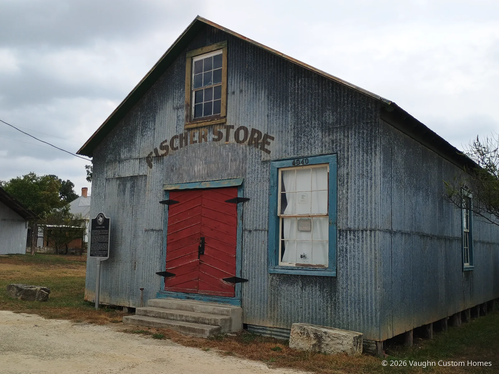 The Fischer Store, a corrugated-metal building with its name painted across the gable, red double doors and a blue-trimmed window, with a Texas Historical Commission marker on a post beside it