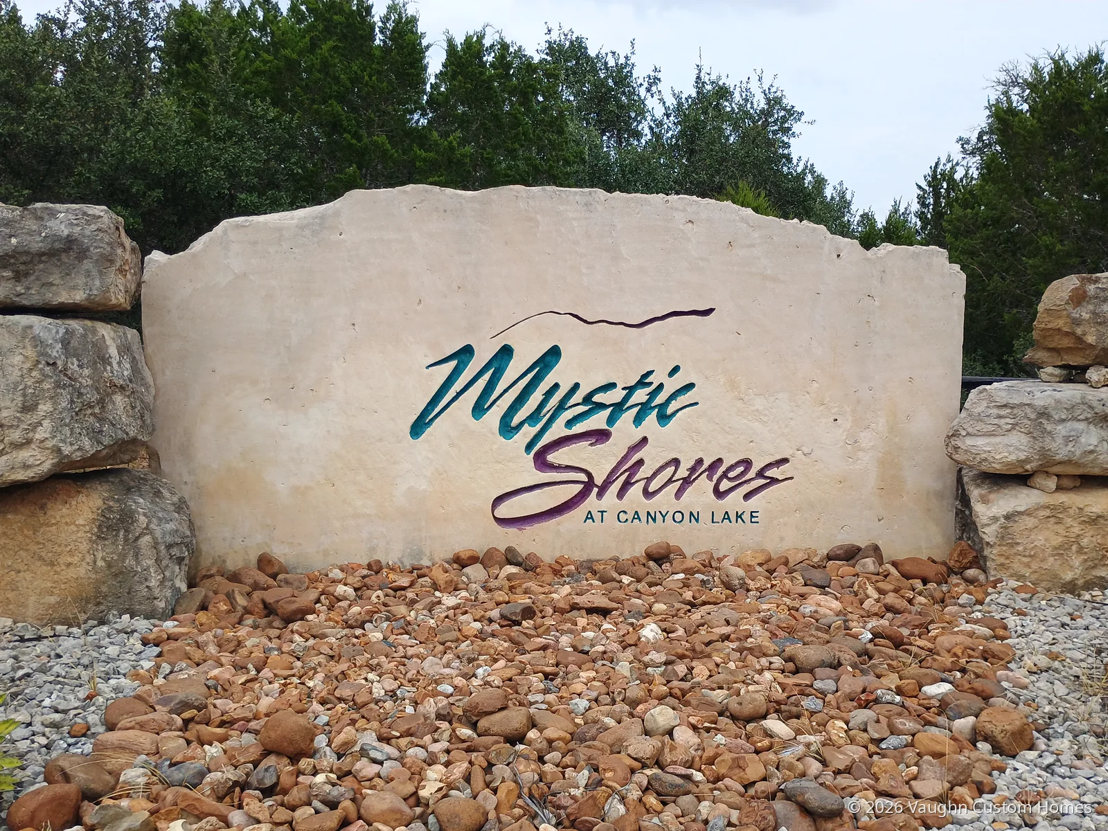 Cream limestone entrance sign reading “Mystic Shores at Canyon Lake” in teal and purple script, set between stacked boulders with a bed of river rock in front and oak trees behind