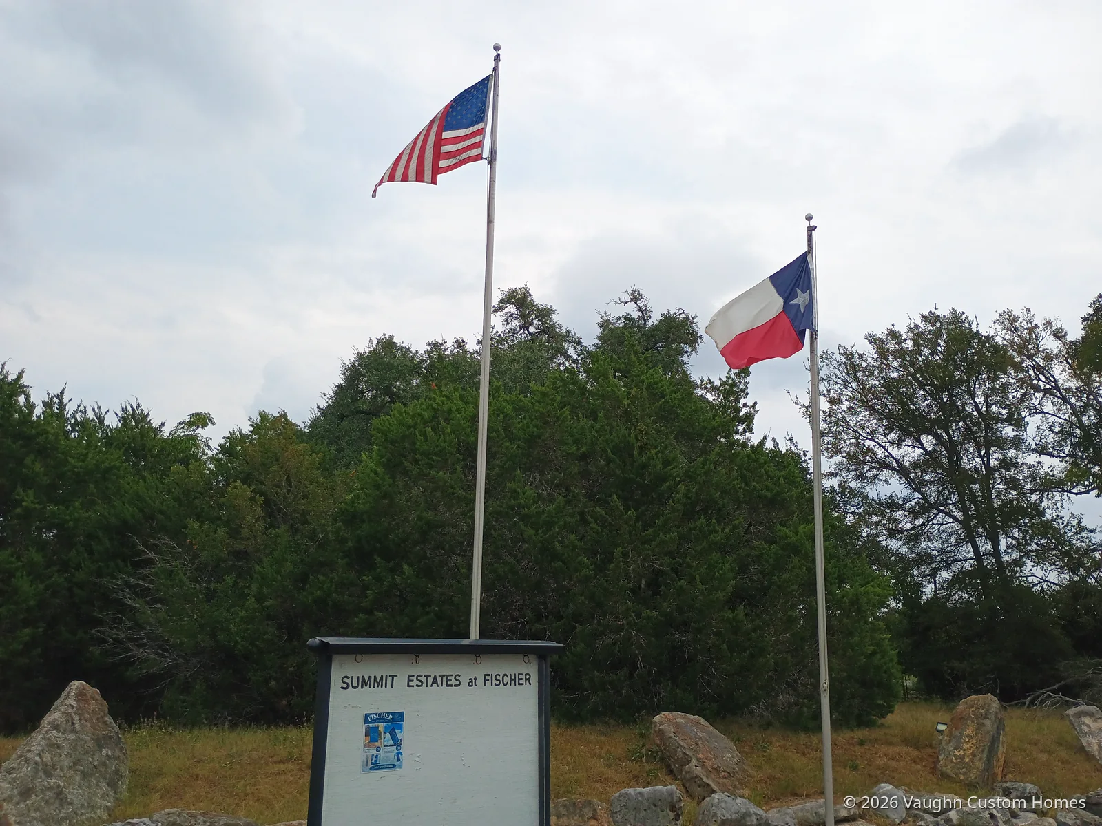 A framed community board reading “Summit Estates at Fischer” beneath a U.S. flag and a Texas flag, with cedar and oak trees behind and rocks scattered across the grass