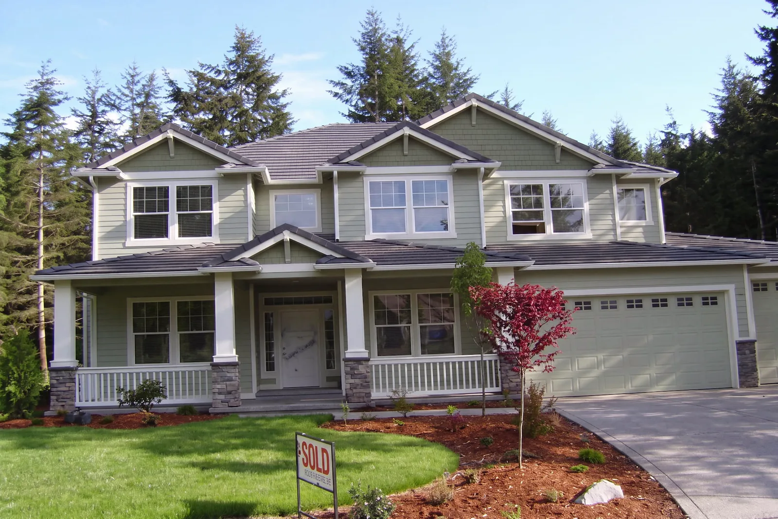 Two-story craftsman home with sage green siding, white trim, a deep covered porch with white railings and stone-based columns, a three-car garage and young maple in the front lawn, surrounded by tall conifers.