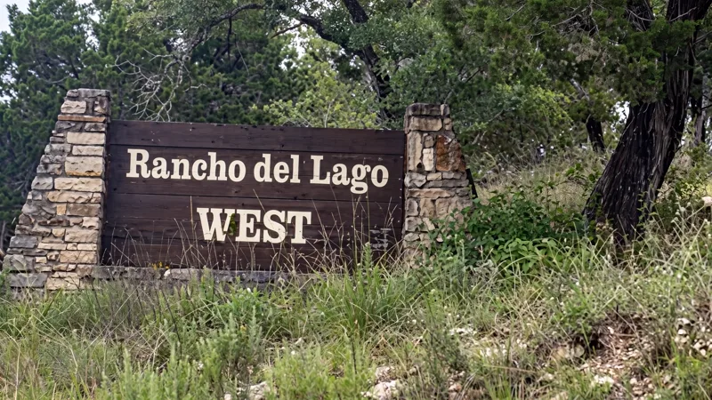 Timber entrance sign reading Rancho del Lago West, set between dry-stacked stone piers among cedar and Hill Country grasses.
